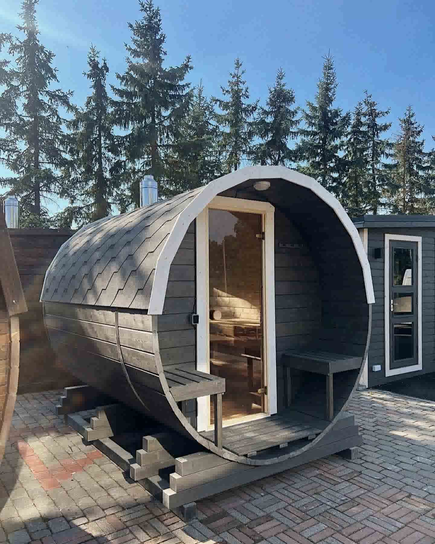 Full side view of a grey-stained barrel sauna with shingle roof and front porch seating area.