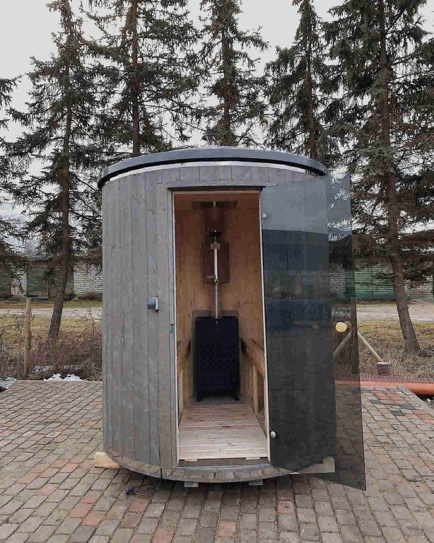 Open door view of a compact vertical sauna showing the internal timber finish and wood-burning stove.