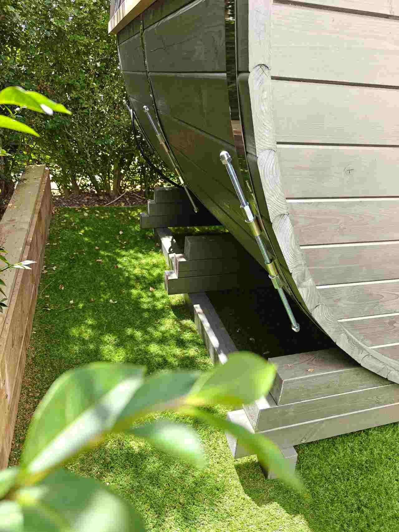 Close-up of the adjustable stainless steel tension bands and bolts securing the grey wood timber planks of a barrel sauna, resting on its wooden support structure in a garden.