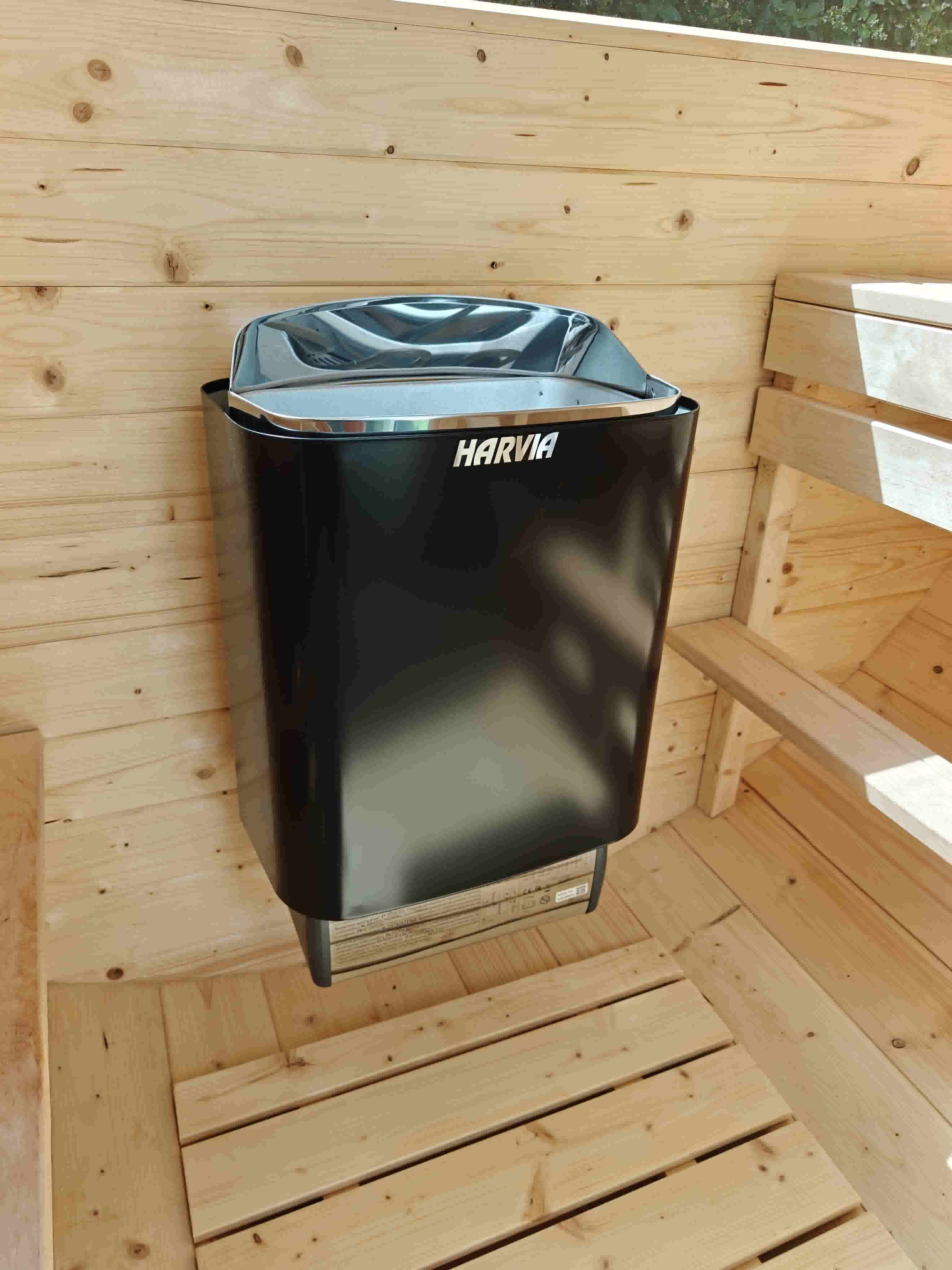 Interior of a barrel sauna featuring a wall-mounted black Harvia electric heater, light-colored natural wood paneling, floor slats, and wooden benches.