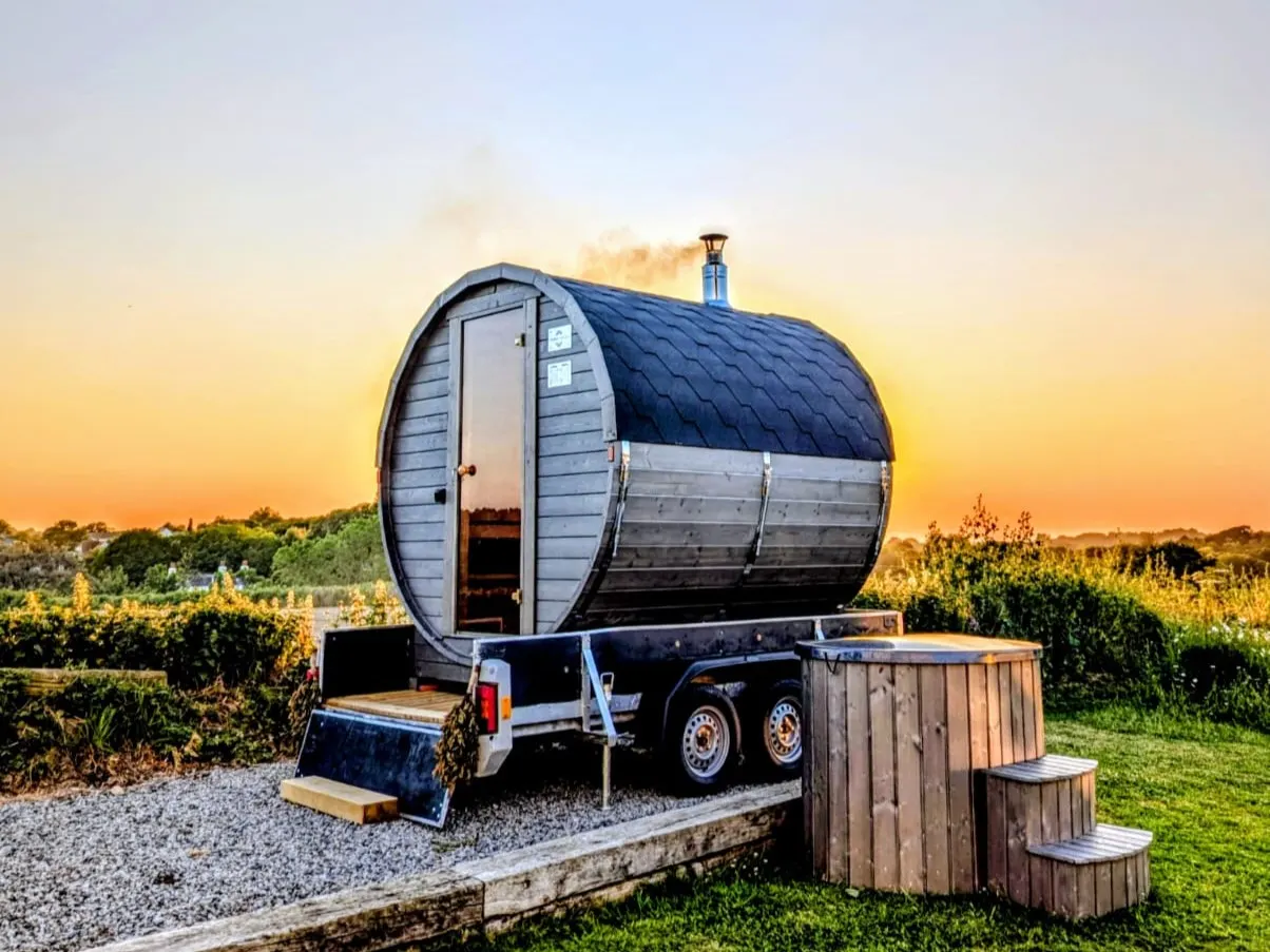 Terra Sauna mobile barrel sauna on a trailer alongside a wooden plunge tub in Guernsey during a scenic sunset.