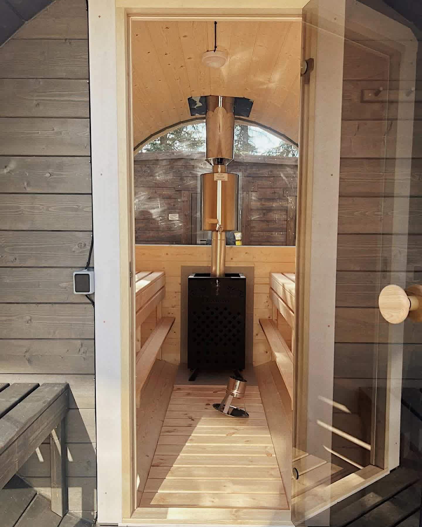 Looking through the open glass door of a Terra Sauna outdoor barrel sauna in Guernsey, featuring a black wood-burning stove with a chimney water tank, natural wood benches, and a half-panoramic glass window.