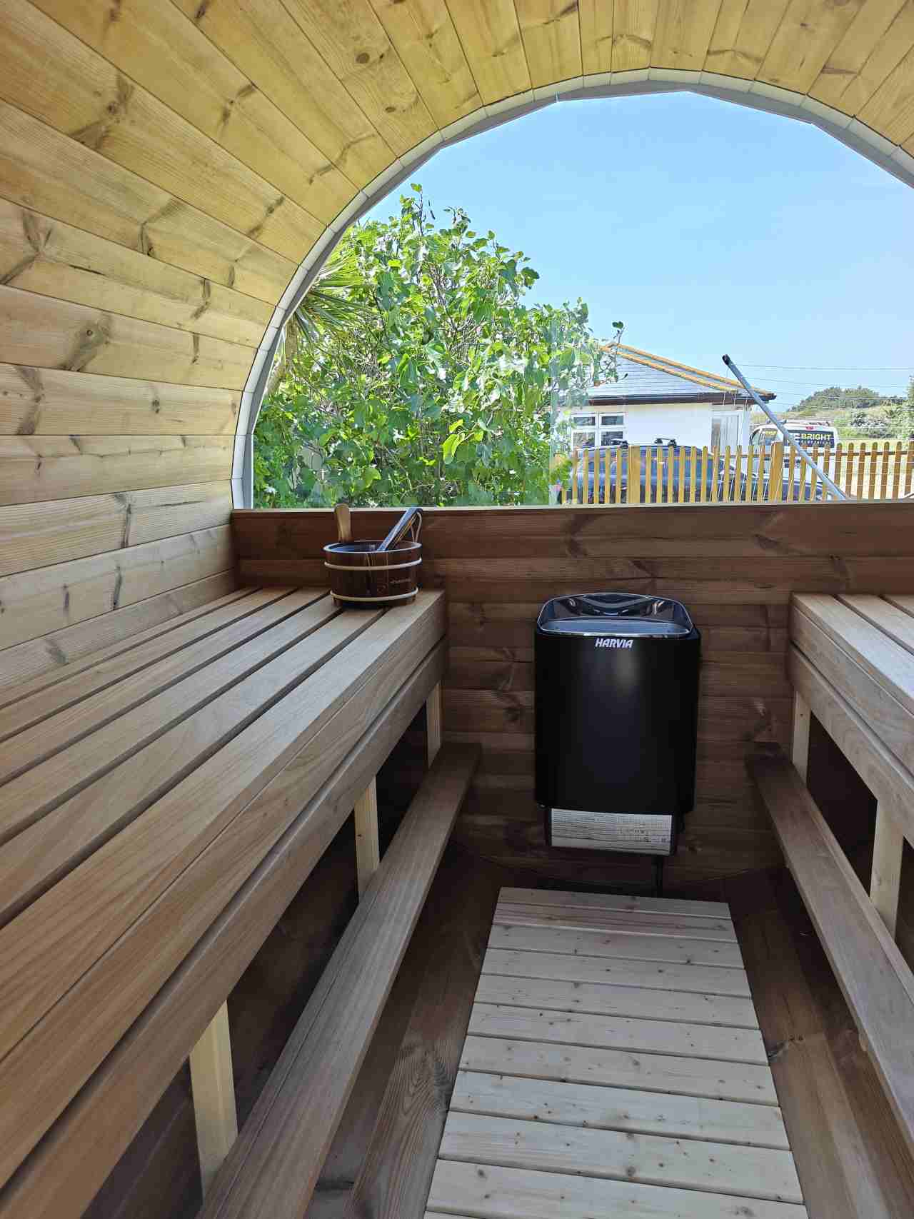 Interior shot from within a Terra Sauna barrel sauna in Guernsey, showing facing wooden benches on the left and right, a central Harvia electric heater, and a traditional wooden bucket and ladle by the arched window, looking out onto a residential scene.