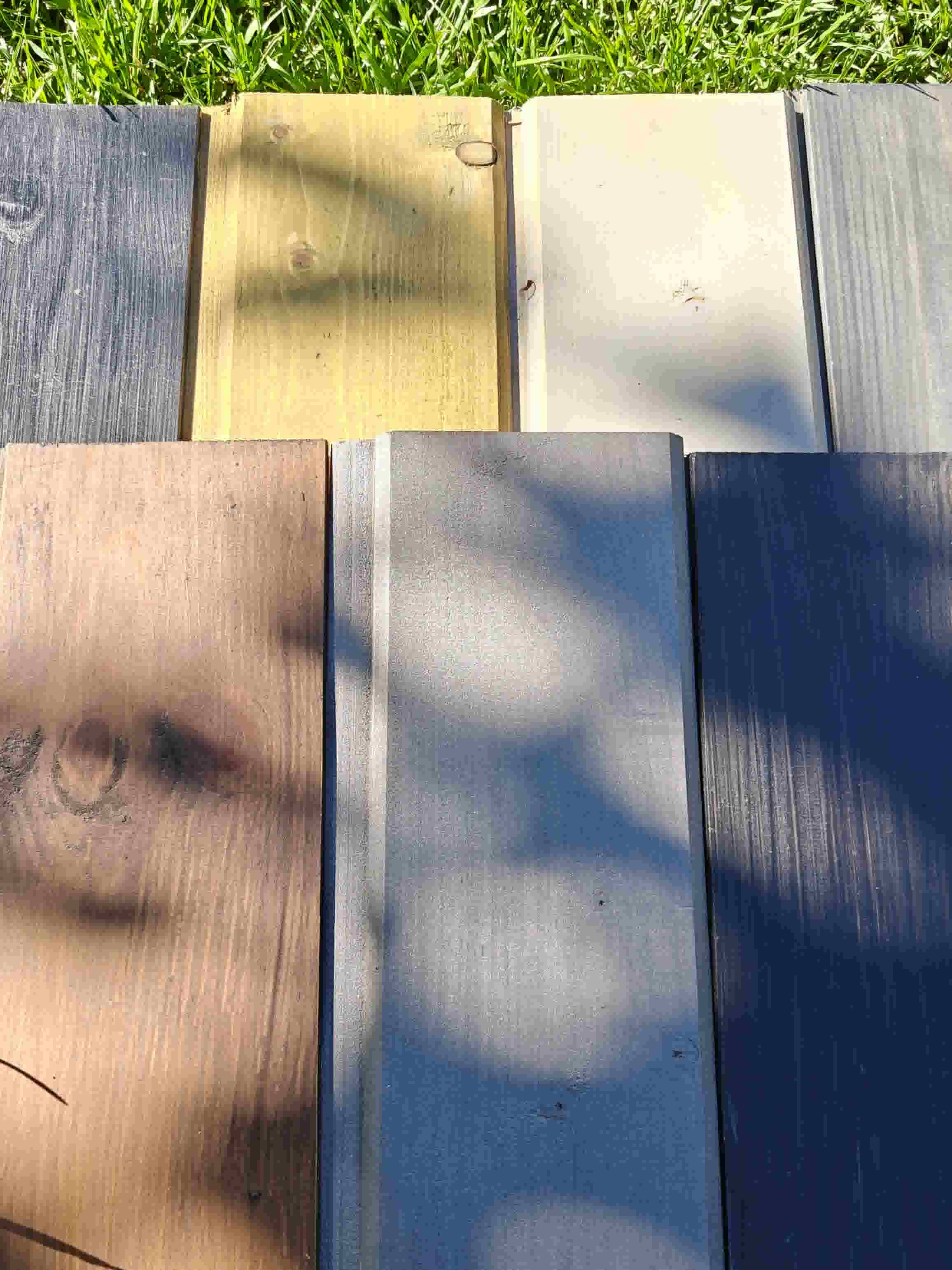 A collection of exterior timber cladding color samples for Terra Sauna builds in Guernsey, arranged side-by-side on green grass, showcasing various wood stains including shades of grey, natural pine, brown, light beige, and charcoal black under natural sunlight.