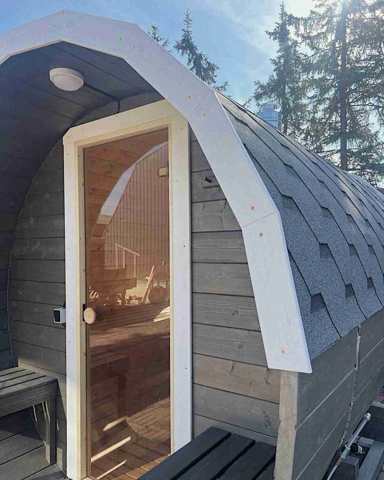 A grey wooden arched barrel sauna by Terra Sauna in Guernsey, showing an external porch with black benches, a glass door, and a shingle roof against a background of trees.