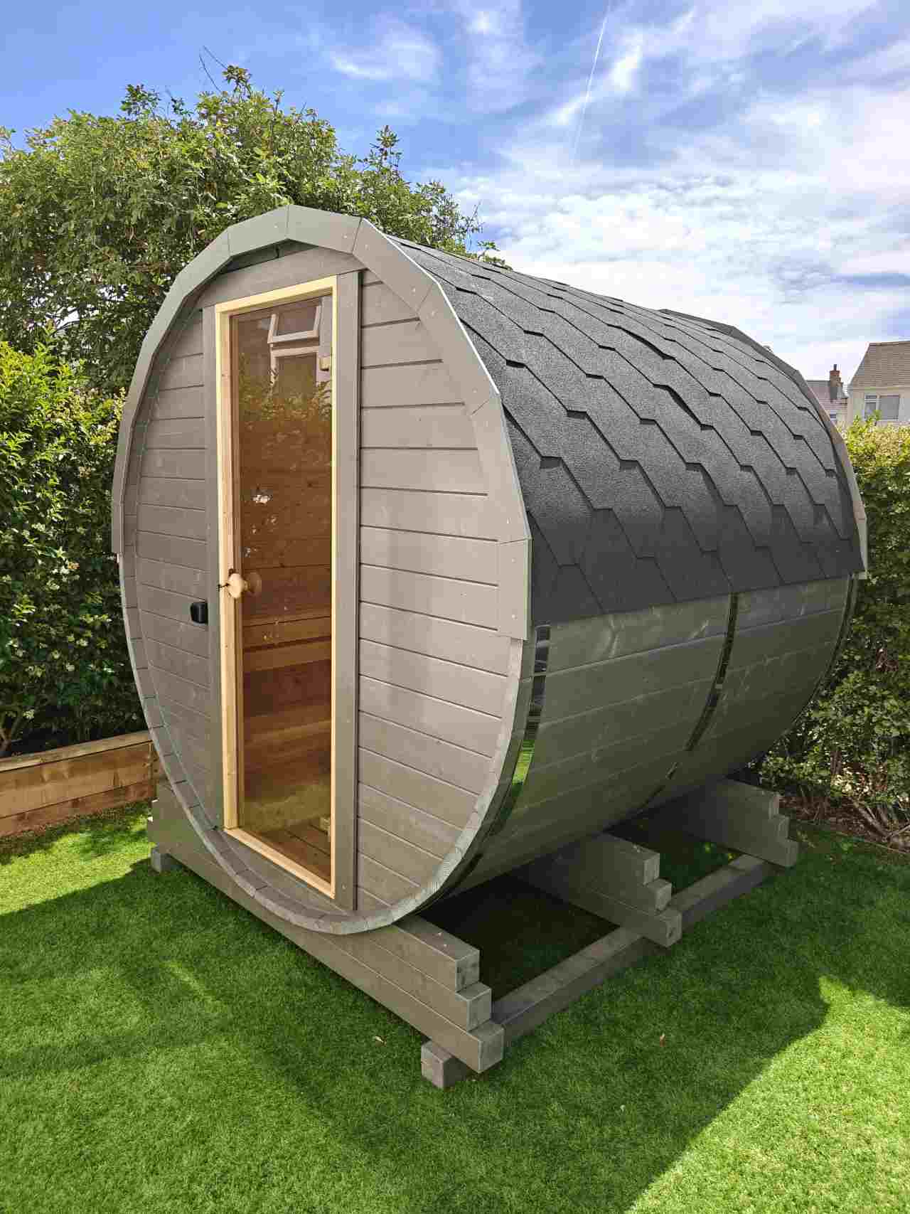 A compact, grey-stained traditional barrel sauna (The Cadus New) with a glass door, situated on a green lawn in a Guernsey garden, featuring black bitumen roof shingles and a sturdy grey support base.