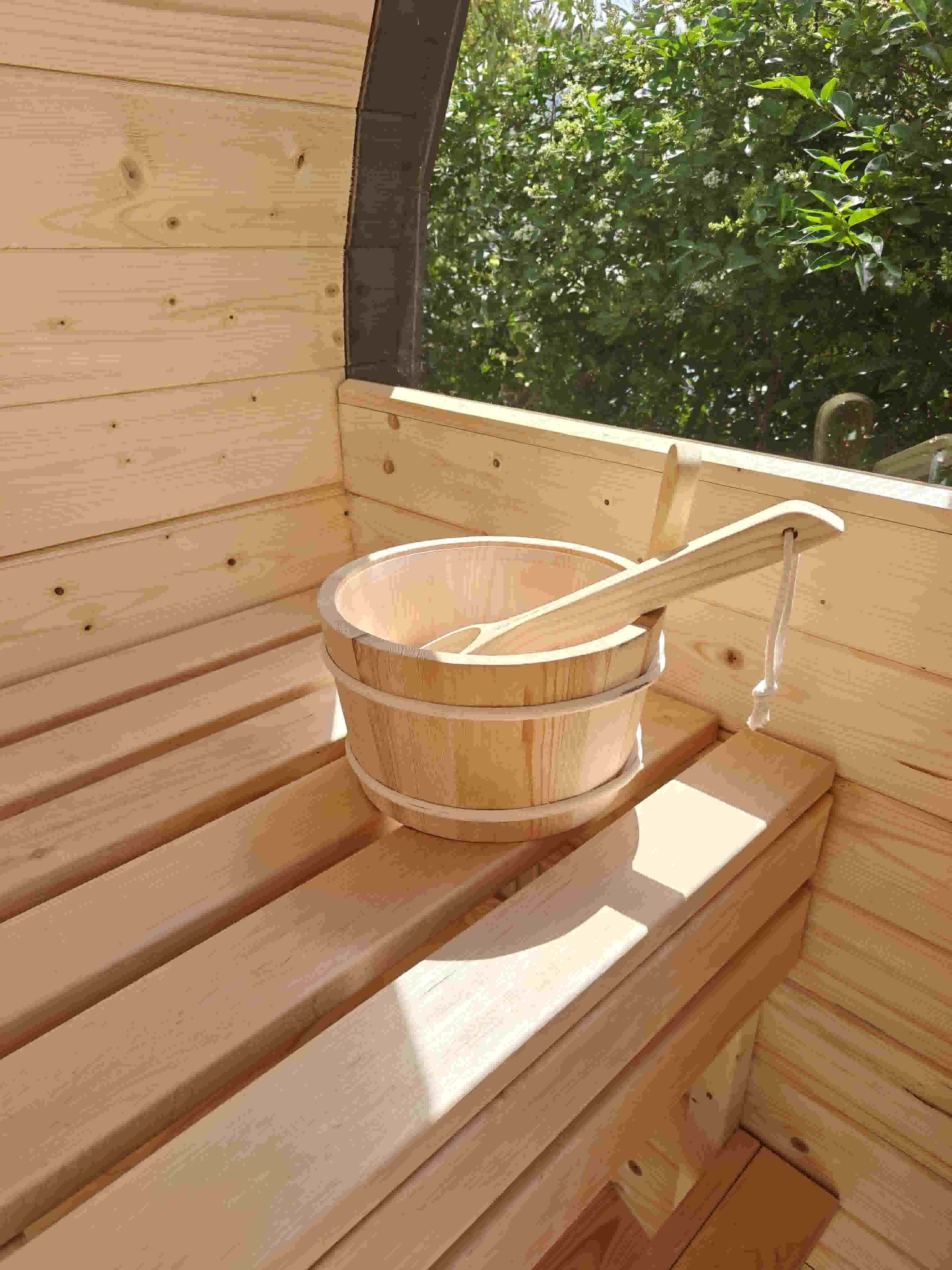 A traditional wooden sauna bucket and ladle sitting on the light wood benches inside a barrel sauna, with natural light coming through a nearby window.